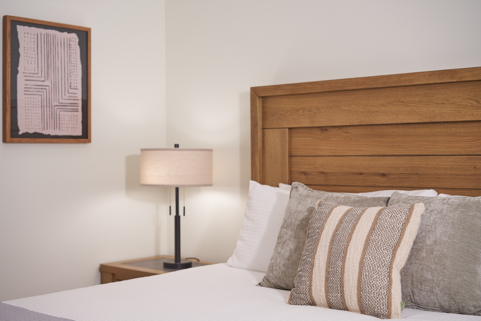 Close view of a finished bedroom with a wood headboard, layered neutral bedding, and a bedside lamp